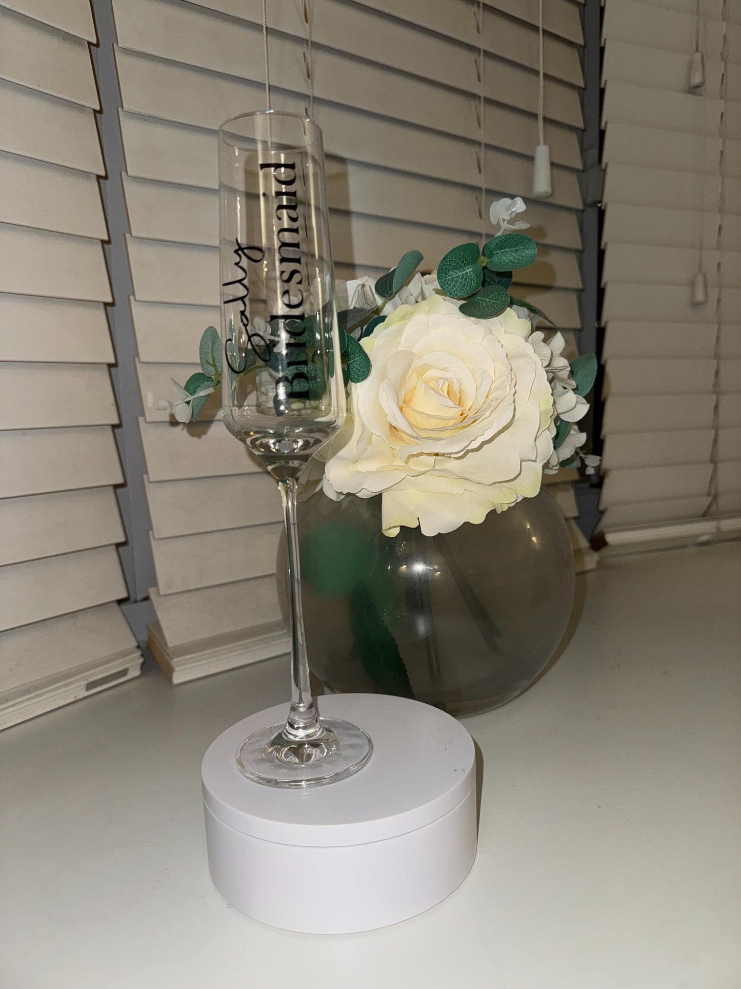Champagne flute with 'Bridesmaid' text next to a white rose in a vase on a plain surface.